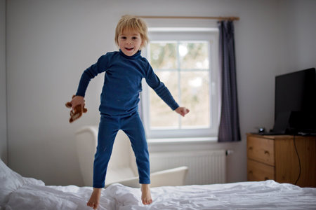 Happy toddler child, blond boy with blue pajama, jumping on the bed, holding little toyの写真素材