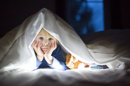 Adorable blond toddler child, cute boy, reading little book with little toy next to him under the duvet in bedの写真素材