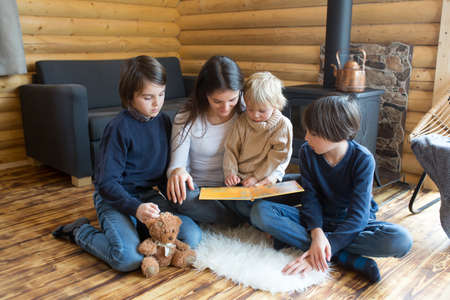 Mother and her three children in a little fancy wooden cottage, reading a book, drinking teaand...