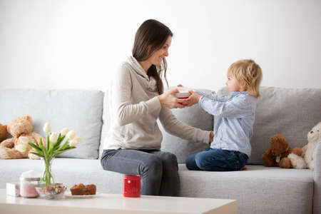 Beautiful blond boy, giving mother flowers and box with little gift for mothers day at sunny living...