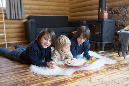Three sweet children, siblings lying on the floor in little fancy wooden cottage, reading a book, drinking teaand enjoying cozy atmosphereの写真素材