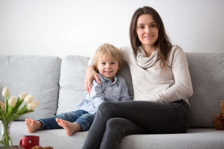 Beautiful blond boy, giving mother flowers and box with little gift for mothers day at sunny living roomの写真素材