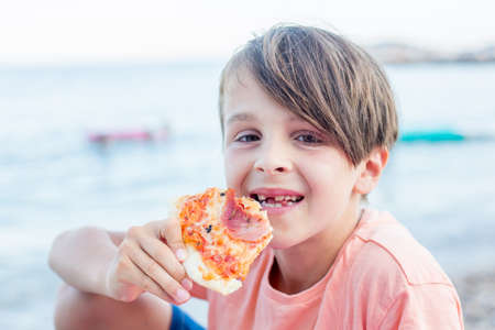 Sweet toddler child, eating pizza on the beach, having fun, smiling happily, kid enjoying dinner at...