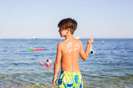 Child, preteen boy with sun protection cream on his back on the beach, holding inflatable ringの写真素材