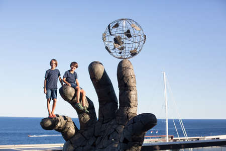 Two children, standing on a art statue of a hand with globe in the center of Monaco, metal hand as art productの写真素材
