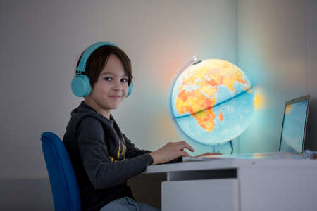 Beautiful child with earphones using laptop while studying at desk at home during coronavirus...