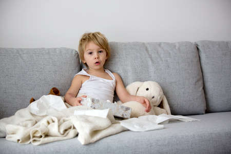 Blond toddler child, wiping his nose in a tissue, sneezing and coughing at home on a sofa