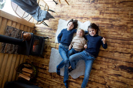Three sweet children, siblings lying on the floor in little fancy wooden cottage, reading a book, drinking teaand enjoying cozy atmosphereの写真素材