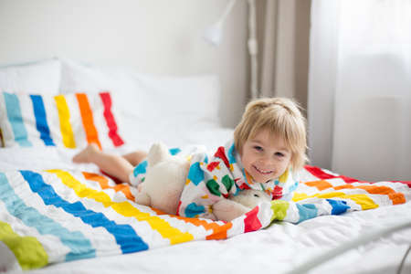 Happy toddler child, blond boy with colorful bathrobe, sitting in bed with stuffed toy after bath,...