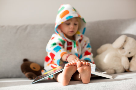 Happy toddler child, blond boy, sitting on the couch with stuffed toy after bath, reading a book, smiling, colorful bathrobeの写真素材