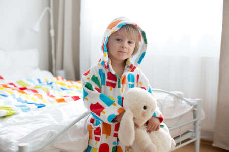 Happy toddler child, blond boy with colorful bathrobe, sitting in bed with stuffed toy after bath, smiling, colorful beddingの写真素材