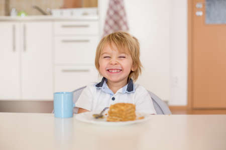 Cute preschool boy, blond child, eating piece of cake and drinking milk at home, happily smilingの写真素材