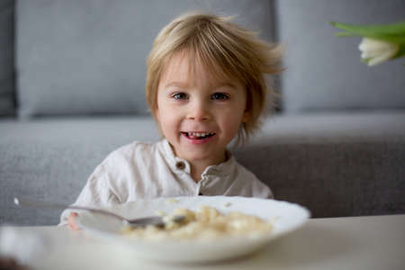 Cute toddler boy, eating pasta with white cheese at home for lunch