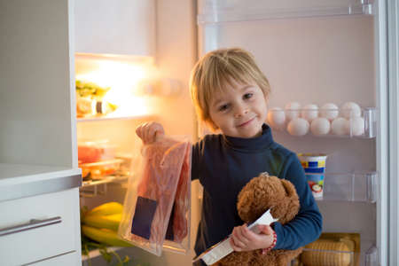 Cute toddler blond child, opening the fridge doo rand taking out smoked meat, prosciutto, healthy food in the refrigeratorの写真素材