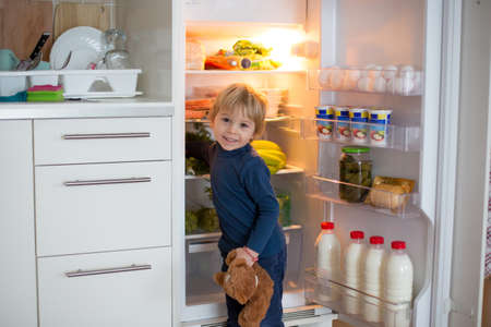 Cute toddler blond child, opening the fridge door and taking fruits adn vegetables and healthy food from the refrigeratorの写真素材