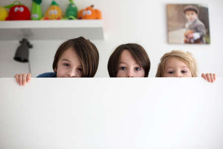 Cute children, three boy brothers, hiding in bed, only eyes peeping out above the wooden board in the morningの写真素材