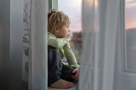 Blond toddler child, sitting on the window on sunset, looking outの写真素材