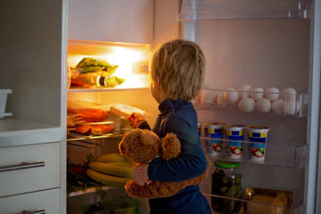 Cute toddler blond child, opening the fridge door and taking fruits adn vegetables and healthy food from the refrigeratorの写真素材