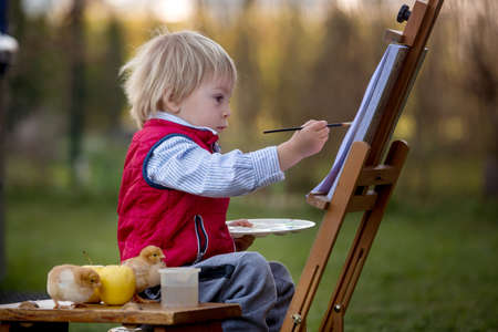 Sweet toddler child, painting eggs in garden with little chicks running around him on sunsetの写真素材