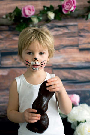Little toddler boy, child with rabbit painted on the face, eating chocolate bunny at home