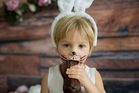 Little toddler boy, child with rabbit painted on the face, eating chocolate bunny at homeの写真素材