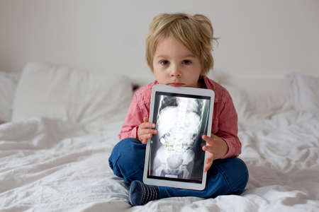 Toddler child, blond boy, holding x-ray picture on tablet of child body with swallowed magnet showing, child swallow dangerous objectの写真素材