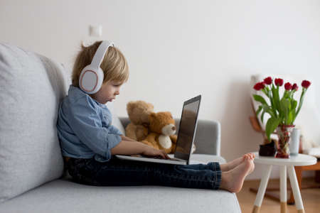 Sweet blond child, boy in casual clothes, woring on computed at home, with headphones and phoneの写真素材