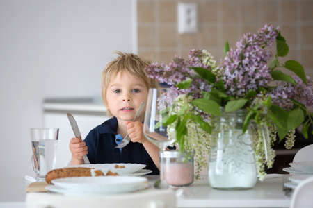 Close portrait of a beautiful blond child, toddler boy with vase with lilacs at home, sitting on the table, ready for lunchの写真素材