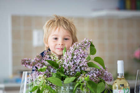 Close portrait of a beautiful blond child, toddler boy with vase with lilacs at home, sitting on the table, ready for lunchの写真素材