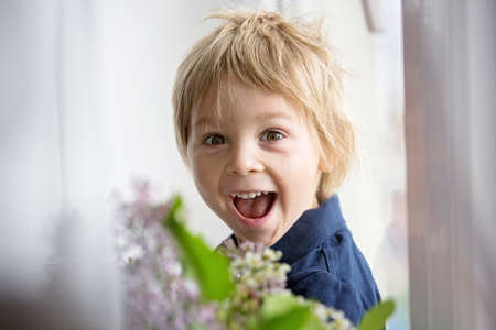 Close portrait of a beautiful blond child, toddler boy with vase with lilacs at home, playing on...