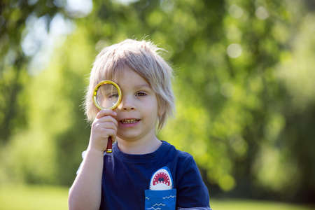 Cute toddler child, blond boy, playing with magnifying glass and drawing in a picture book in the park, summertimeの写真素材