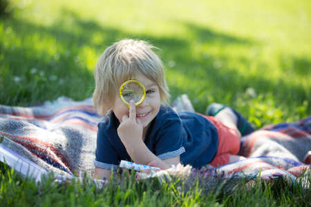 Cute toddler child, blond boy, playing with magnifying glass and drawing in a picture book in the park, summertimeの写真素材
