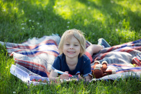 Cute toddler child, blond boy, playing with magnifying glass and drawing in a picture book in the park, summertimeの写真素材