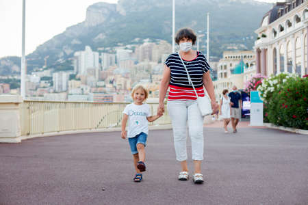 Grandmother and toddler child, walking hand in hand in the center of Monaco, summer 2021, medical masks required everywhereの写真素材