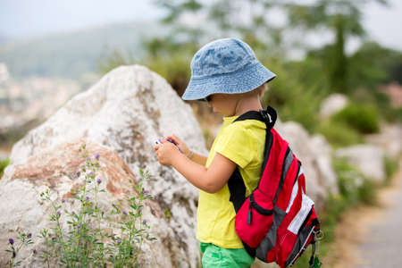 Cute preschool boy with backpack, walking in a mountain on a cloudy day, taking pictures of natureの写真素材