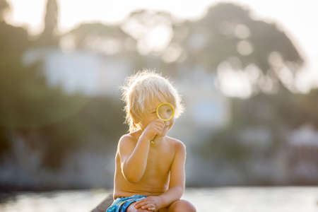 Cute child, boy, using magnifying glass to examine seashells on the beach, child in water in the seaの写真素材