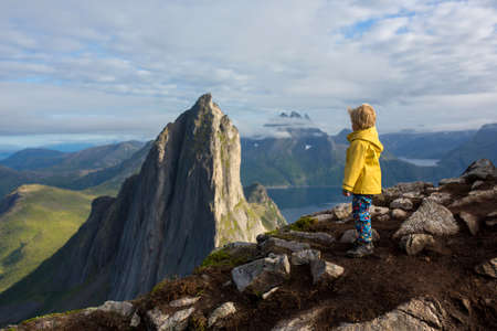 Cute toddler child in yellow raincoat, standing backwards on a rock and looking over Segla mountain on Senja island, North Norway. Amazing beautiful landscape and splendid natureの写真素材