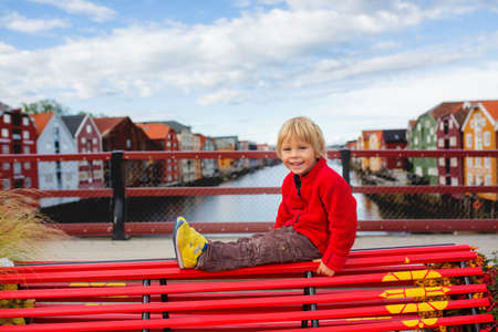 Cute toddler blond child, visiting Trondheim, Norway during the summer, sitting on a red bench, enjoying the cityの写真素材