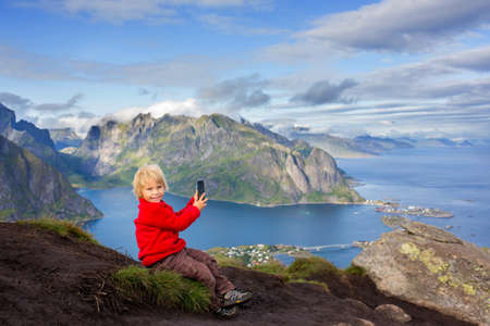 Cute child, standing on top of the mountains and looking down on Reine after climbing Reinebringen treeking path with lots of stairs, taking pictures with cellphoneの写真素材