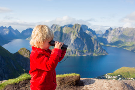 Cute child, standing on top of the mountains and looking down on Reine after climbing Reinebringen treeking path with lots of stairs, using binoculars to look at the viewの写真素材
