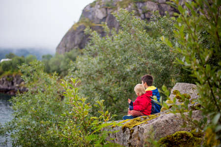 Beautiful children, cute boys, sitting on a rock on the edge of a fjord in Lofoten, enjoying the viewの写真素材
