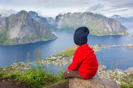 Cute child, standing on top of the mountains and looking down on Reine after climbing Reinebringen treeking path with lots of stairsの写真素材