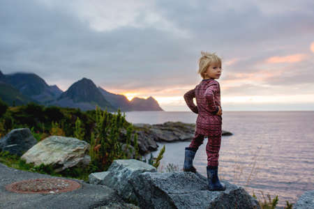 Cute child, enjoying amazing view from a rock in Husoy on Senja island, North Norway. Amazing beautiful landscape and splendid nature in scandinavian country Norwayの写真素材