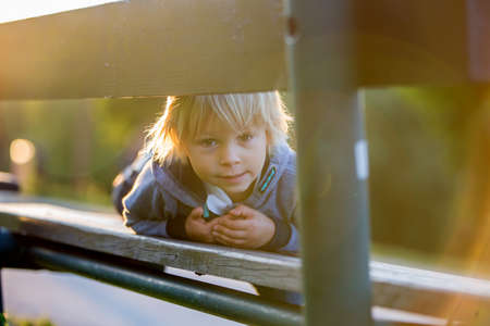 Close portrait of a cute blond child, boy, lying on bench in the park on sunset, summertime