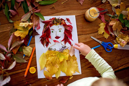 Toddler blond child, creating woman portrait of leaves in living room, applying leaves using glue, autumntimeの写真素材