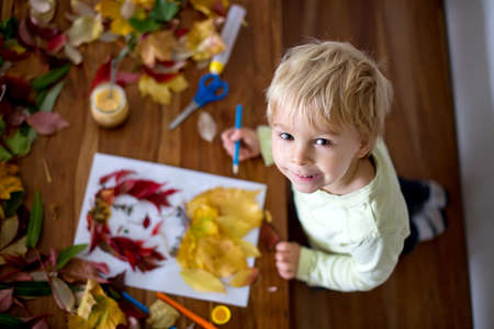 Toddler blond child, creating woman portrait of leaves in living room, applying leaves using glue, autumntimeの写真素材