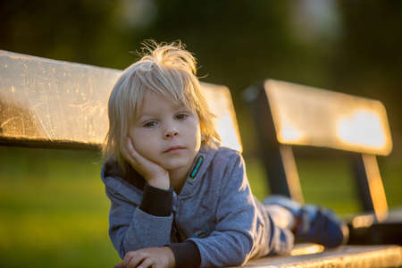 Close portrait of a cute blond child, boy, lying on bench in the park on sunset, summertimeの写真素材