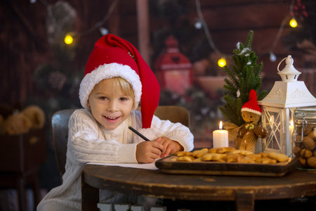 Cute toddler child, boy on Christmas, writing letter to Santa Claus and eating cookies at home, cozy wooden cottageの写真素材