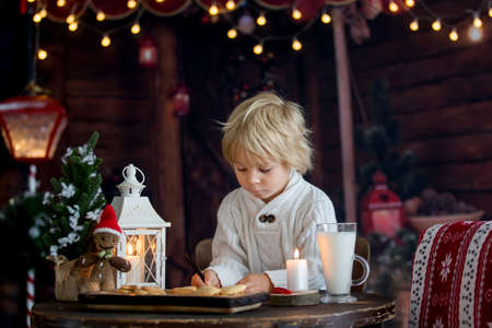 Beautiful toddler child, blond boy, writing letter to Santa Claus and eating cookies at home, cozy atmosphere on Christmasの写真素材