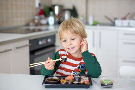 Cute blond child Eat Sushi Rolls At Home. Happy boy ready for eating sushi, sitting in a sunny kitchenの写真素材